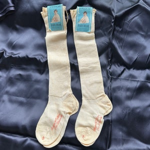 May include: Two pairs of off-white knee-high stockings with the brand name "American Baby Hose" printed on the tags. The stockings are on a dark blue fabric background.