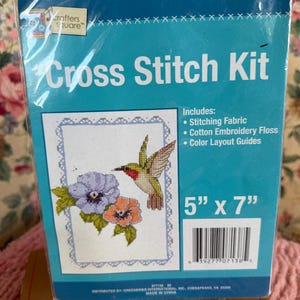 Puede incluir: Un kit de punto de cruz con un diseño de flores y un colibrí. El kit incluye tela de bordado, hilo de bordar de algodón y guías de color. El paquete está etiquetado como "Cross Stitch Kit" y mide 12,7 cm x 17,8 cm.