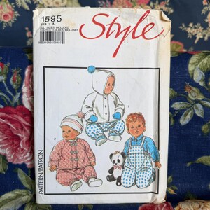 Puede incluir: Un sobre de papel para patrón de costura con la etiqueta Style 1595, que presenta ilustraciones de tres bebés con varios atuendos, incluyendo una chaqueta con capucha, un conjunto acolchado y un overol de lunares, con un pequeño panda incluido.
