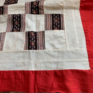 May include: A white patchwork quilt with a red border. The quilt is made of squares of different fabrics, some with floral patterns.