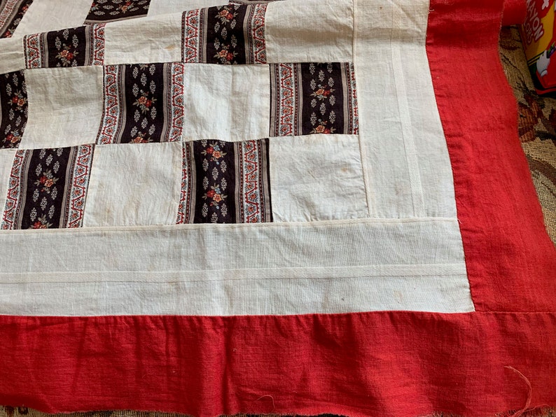 May include: A white patchwork quilt with a red border. The quilt is made of squares of different fabrics, some with floral patterns.