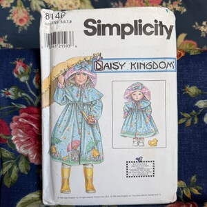 May include: A Simplicity Daisy Kingdom sewing pattern envelope, number 8146, featuring an illustration of a child and a doll wearing a light blue raincoat with a colorful umbrella print, a matching bonnet, and yellow rain boots, with text included.