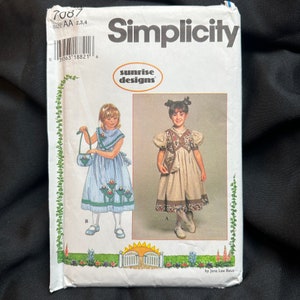 May include: Simplicity sewing pattern 7087 for girls' dresses. The pattern includes two designs: a blue and white striped dress with a flower applique and a brown and white plaid dress with a floral yoke. The pattern is from the Sunrise Designs collection.