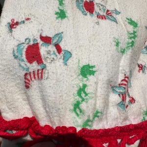 May include: White fabric with a repeating pattern of red, blue, and green figures. The edge is trimmed with a red, crocheted border. The fabric appears to be a towel or similar absorbent material.