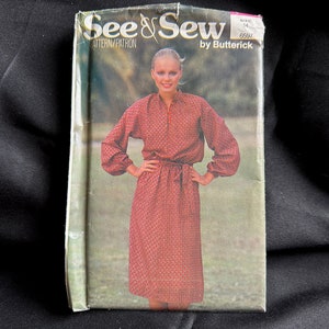 Puede incluir: Un patrón de costura vintage para un vestido, con una mujer que lleva un vestido estampado en rojo y blanco con mangas largas y un cinturón de lazo. El patrón está etiquetado como "See & Sew" de Butterick, talla 14, 6597.