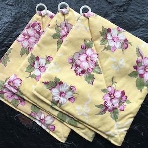 Könnte beinhalten: Drei gelbe Topflappen aus Stoff mit einem Blumenmuster in den Farben Rosa, Weiß und Grün. Die Topflappen haben weiße Schlaufen zum Aufhängen.