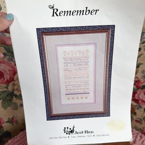 May include: A framed cross-stitch sampler with the word "Remember" at the top. The sampler features a poem with floral accents in shades of pink and lavender. The frame has a dark blue and burgundy floral pattern.