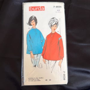 May include: A vintage Burda sewing pattern packet featuring illustrations of two blouses. One blouse is blue with a criss-cross neckline, and the other is red with a tie-neck. The packet has text in German, French, and English.