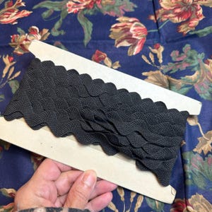 May include: A roll of black scalloped trim on a wooden spool. The trim has a textured, woven appearance and is displayed against a floral patterned fabric background.