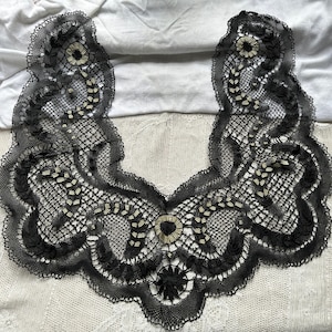 May include: Black lace collar with a delicate floral design and gold accents. The collar is made of intricate crocheted lace and has a scalloped edge.