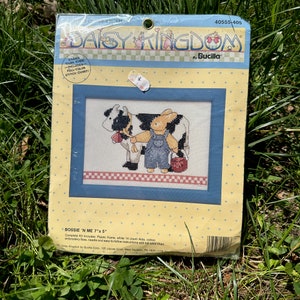 May include: A cross-stitch kit featuring a bunny and a cow. The kit includes a plastic frame, white 14 count Aida, cotton embroidery floss, a needle, and easy-to-follow instructions with a full-color chart. The kit is titled "Bossie 'N Me" and measures 7 inches by 5 inches.