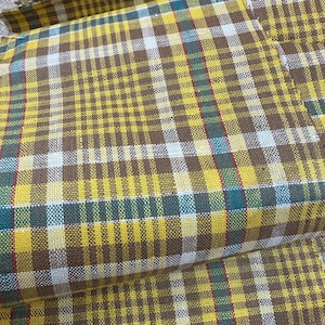 May include: Close-up of a folded fabric with a plaid pattern. The pattern features a grid of yellow, brown, white, teal, and red lines. The fabric appears to be woven, with a slightly textured surface.