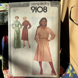 Peut inclure: Patron de couture Simplicity 9108, taille 14 Miss. L'enveloppe du patron présente des illustrations de trois modèles de robes. L'image principale montre une femme dans une robe rose clair avec une taille nouée. L'enveloppe est blanc cassé avec du texte noir.