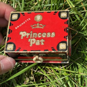 May include: A vintage Princess Pat box, red with gold trim and lettering. The box features the text "Princess Pat" in a decorative font, with "Brunette" at the top. The box is rectangular with a small clasp.