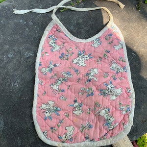 Pode incluir: Um babador de bebê acolchoado rosa com uma borda branca e um padrão de coelhos brancos e flores azuis.