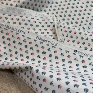 May include: White fabric with a repeating pattern of small pink flowers and blue leaves. The fabric has the text "Laura Ashley MCM LXXXIX" and "Printed in U.K." printed on it.