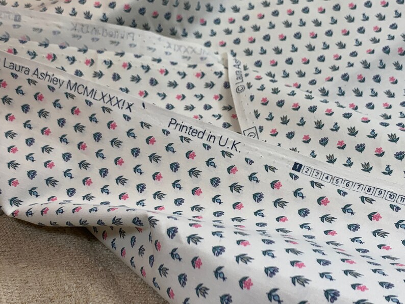 May include: White fabric with a repeating pattern of small pink flowers and blue leaves. The fabric has the text "Laura Ashley MCM LXXXIX" and "Printed in U.K." printed on it.