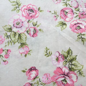 May include: White fabric with a repeating pattern of pink roses and green leaves. The floral design features various sizes of roses, with some in full bloom and others as buds. The fabric has a slightly textured appearance.
