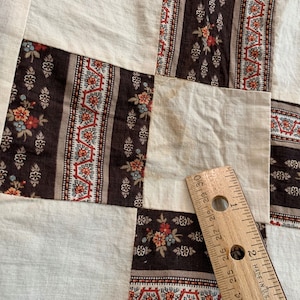 May include: A close-up of a patchwork quilt with a brown, white, and red floral pattern. A ruler is placed on the quilt to show the scale.