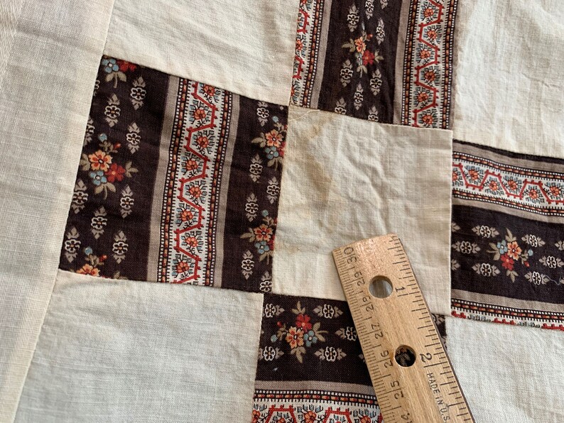 May include: A close-up of a patchwork quilt with a brown, white, and red floral pattern. A ruler is placed on the quilt to show the scale.