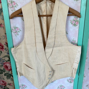 May include: A cream-colored vest with a pinstripe pattern and a shawl collar. The vest has three buttons and two pockets.
