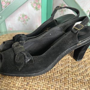 Può includere: Un paio di scarpe con tacco slingback nere in camoscio con punta aperta. Ogni scarpa presenta un dettaglio a fiocco sulla tomaia e una chiusura con fibbia sul cinturino posteriore. I tacchi sono alti circa 7.6 cm.