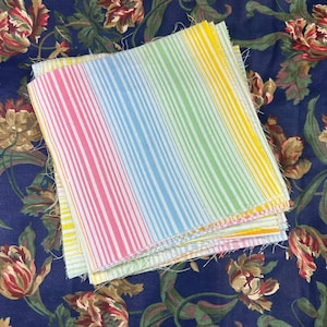 May include: A stack of colorful fabric squares with vertical stripes in pink, blue, green, and yellow. The fabric is set against a dark blue floral background.