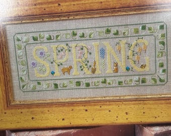 May include: Cross-stitch artwork in a wooden frame. The word "SPRING" is stitched in yellow, with floral and animal motifs. The frame is a light brown color, and the background is a neutral beige.