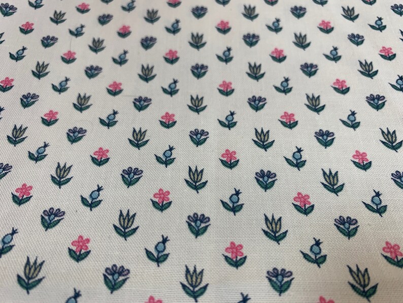 May include: White fabric with a repeating pattern of small pink and blue flowers with green stems. The flowers are stylized and have a simple design.