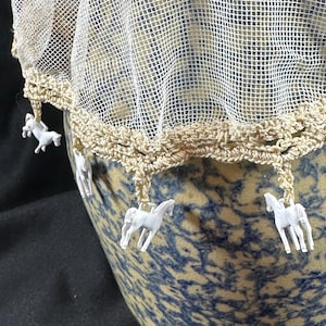 May include: A decorative, off-white net cover with a crocheted edge, adorned with small white horse figurines. The cover is draped over a blue and tan ceramic pot, creating a rustic, country aesthetic.