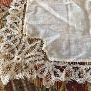 May include: A white linen handkerchief with cream-coloured lace edging. The lace features floral and geometric patterns, adding a delicate, vintage aesthetic. The handkerchief is square-shaped, with a detailed lace corner.