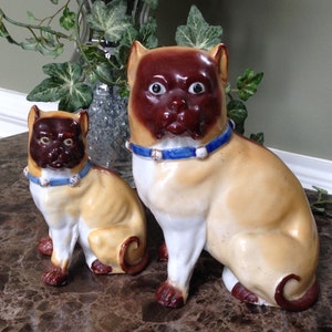 May include: Two ceramic figurines of pugs, one larger than the other. Both are sitting with their tails curled. The larger pug is brown and white with a blue collar. The smaller pug is tan and white with a blue collar.