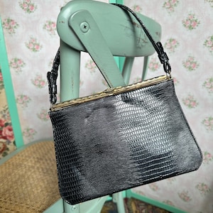 May include: A vintage black handbag with a textured, reptile-like pattern. The bag features a gold-toned clasp and a thin black strap. It is hanging on a light green chair with a floral background.