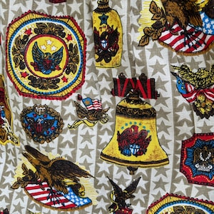 May include: A beige fabric with a repeating pattern of stars, eagles, bells, and flags. The fabric features a patriotic theme with red, white, and blue colours.