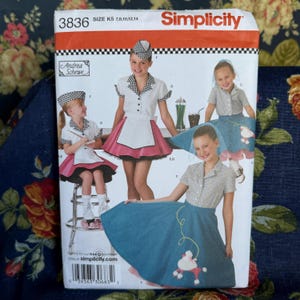 May include: A Simplicity sewing pattern envelope 3836 for size K5 7, 8, 10, 12, 14. The cover features illustrations of outfits including a white collared shirt with a black and white checkered trim, a white apron, and a pink skirt, plus a blue skirt with a poodle applique.