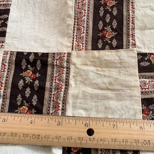 May include: A close-up of a quilt made of different fabric squares. The squares are in a variety of patterns, including floral and geometric designs. The quilt is made of brown, white, and red fabrics. A wooden ruler is placed on top of the quilt, with the numbers 2, 3, 4, 5, 6, 7, 8, and 9 visible.