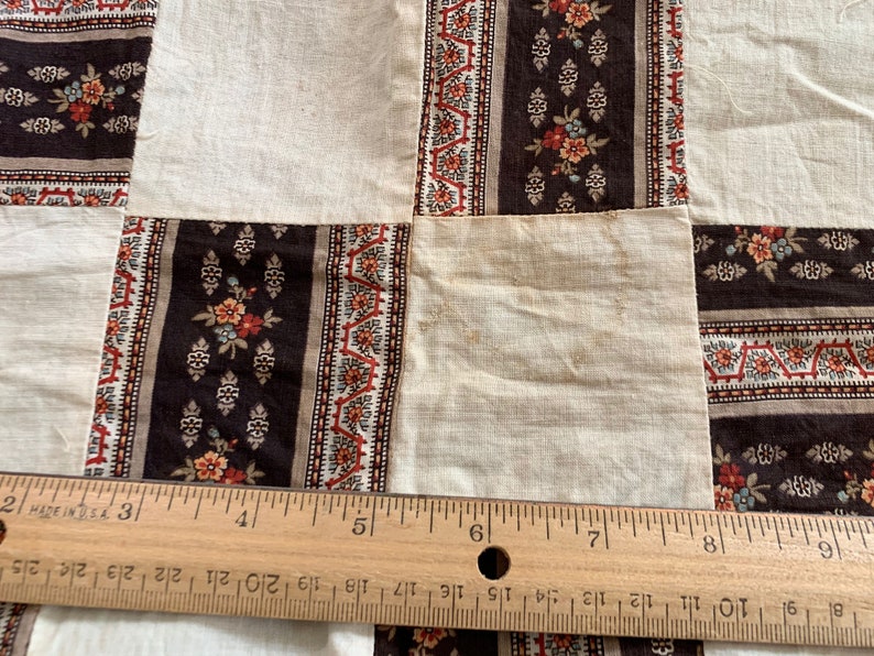 May include: A close-up of a quilt made of different fabric squares. The squares are in a variety of patterns, including floral and geometric designs. The quilt is made of brown, white, and red fabrics. A wooden ruler is placed on top of the quilt, with the numbers 2, 3, 4, 5, 6, 7, 8, and 9 visible.