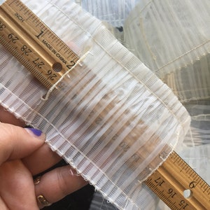 Può includere: Primo piano di un tessuto bianco plissettato, misurato con un righello. Il righello, segnato in centimetri, mostra le dimensioni del tessuto. Il tessuto sembra essere un materiale delicato e trasparente, possibilmente per abbigliamento o progetti artigianali.