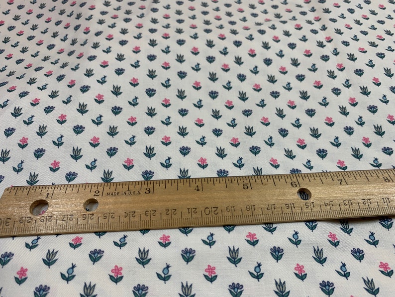 May include: White fabric with a repeating pattern of small pink and blue flowers. A wooden ruler is placed on top of the fabric.