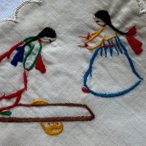 Może przedstawiać: Haftowany biały obrus lniany z dwoma postaciami w kolorowych ubraniach. Postacie to kobieta w niebieskiej sukience i kobieta w czerwonej i zielonej sukience. Kobieta w niebieskiej sukience stoi, a kobieta w czerwonej i zielonej sukience siedzi na brązowej drewnianej ławce.