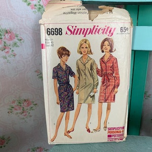 Op de afbeelding: Vintage Simplicity naaipatroon nummer 6698 voor een jurk. Het patroon is voor damesmaten 40 en heeft een borstomvang van 42. De patroonhoes toont drie vrouwen die verschillende versies van de jurk dragen. De jurk is een midi-jurk, met knopen, een kraag en lange mouwen.