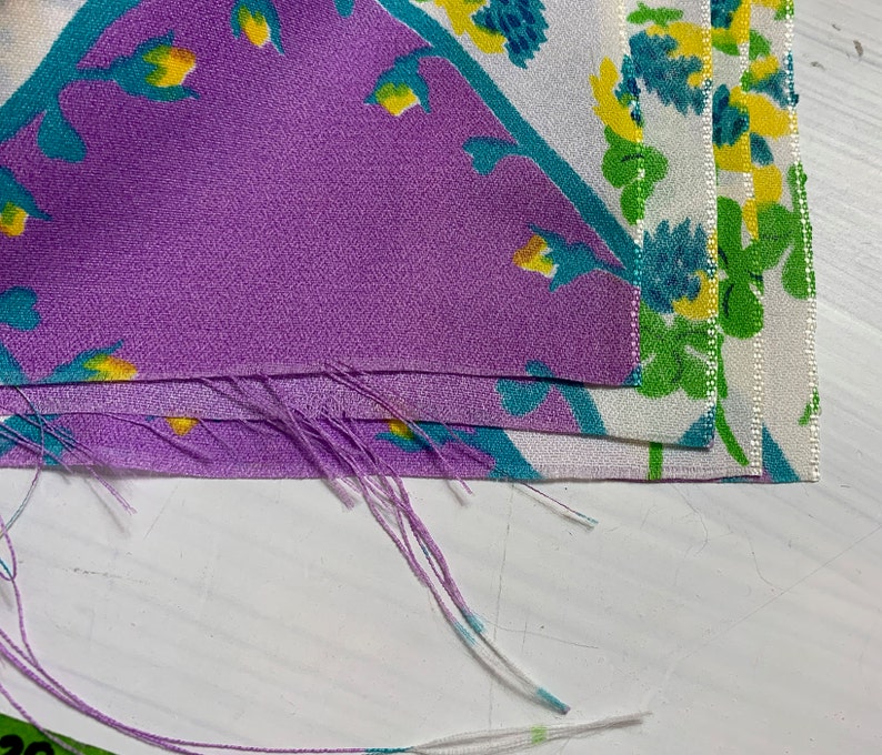Pu&ograve; includere: Primo piano di un mucchio di ritagli di tessuto con un motivo floreale in tonalit&agrave; di viola, bianco, verde e giallo. Il tessuto &egrave; sfilacciato ai bordi.