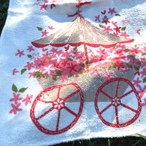May include: White towel with a floral design. The design includes a parasol, flowers, and wagon wheels in pink, red, and green. The towel has a soft, textured appearance, possibly made of cotton or a similar material.