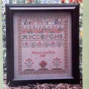 May include: A framed cross-stitch sampler with a brown wooden frame. The sampler features a traditional alphabet, numbers, and a floral design with red flowers. The text "Margaret Jessie Docker. 1835" is stitched in the bottom center of the sampler.