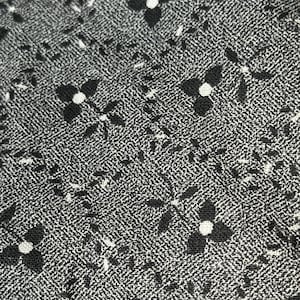 May include: Close-up of a fabric with a repeating pattern of black four-petal flowers with white centers on a textured gray background. Small white dashes and dots fill the spaces between the flowers.