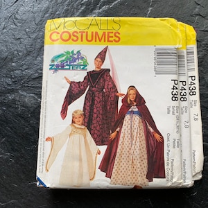 Puede incluir: Tres patrones de costura para disfraces, con un disfraz de bruja morado y negro, una capa roja con capucha y un vestido de princesa blanco. Los patrones están etiquetados como "P438" y son para las tallas 7 y 8.