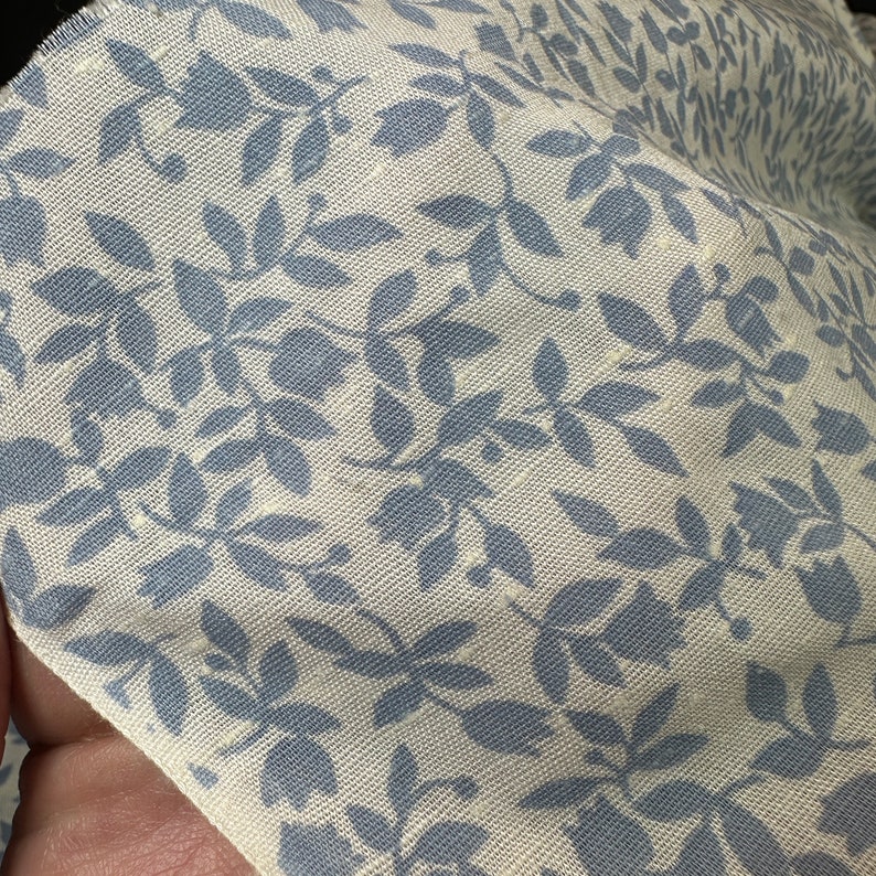 May include: Close-up of a white fabric with a blue floral pattern. The fabric has a subtle texture and the floral design is repeated throughout the fabric.