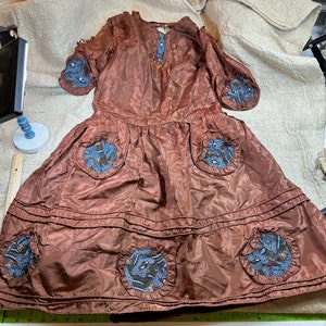 Peut inclure: Une robe vintage marron avec des embellissements en perles bleues et blanches. La robe a un col rond et des manches longues. La jupe est à étages et a un aspect plissé.