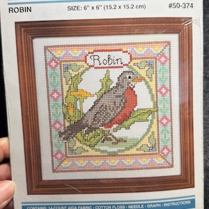 May include: A cross-stitch kit featuring a framed image of a robin bird. The kit is labeled "Weekend Cross Stitch" and includes a 6" x 6" (15.2 x 15.2 cm) design. The kit contains 14-count Aida fabric, cotton floss, a needle, graph, and instructions.