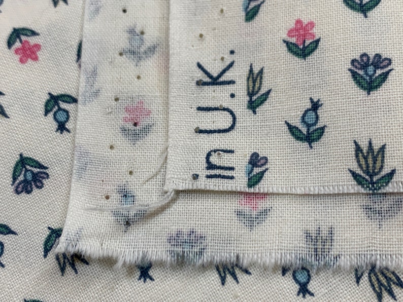 May include: Close-up of two pieces of white fabric with a floral pattern. The fabric has a small, repeating pattern of blue, green, and pink flowers. The top piece of fabric has the word "inuk" printed on it in black.